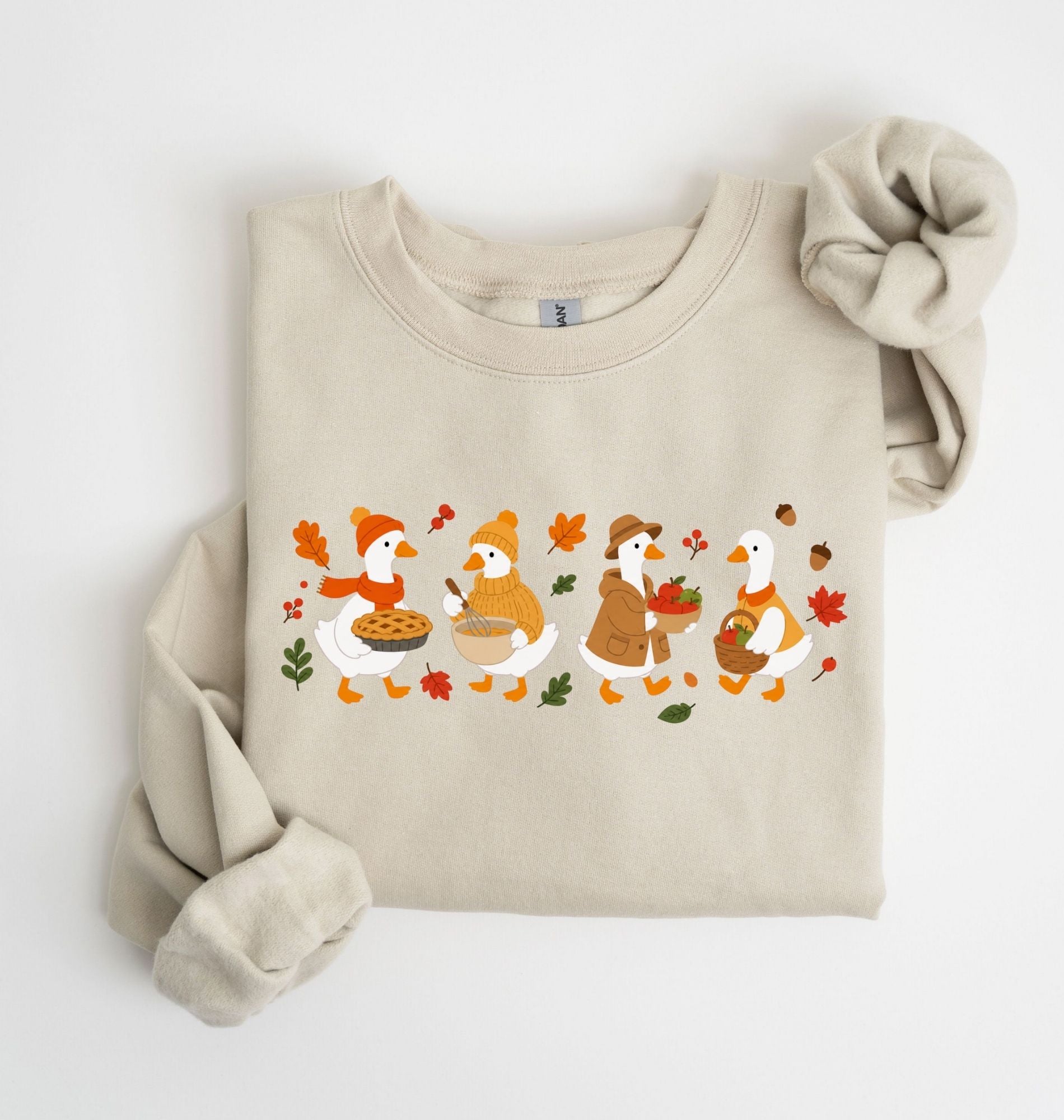 Flat lay of a sand fall sweatshirt featuring four geese baking pie, mixing ingredients and gathering apples on a white background