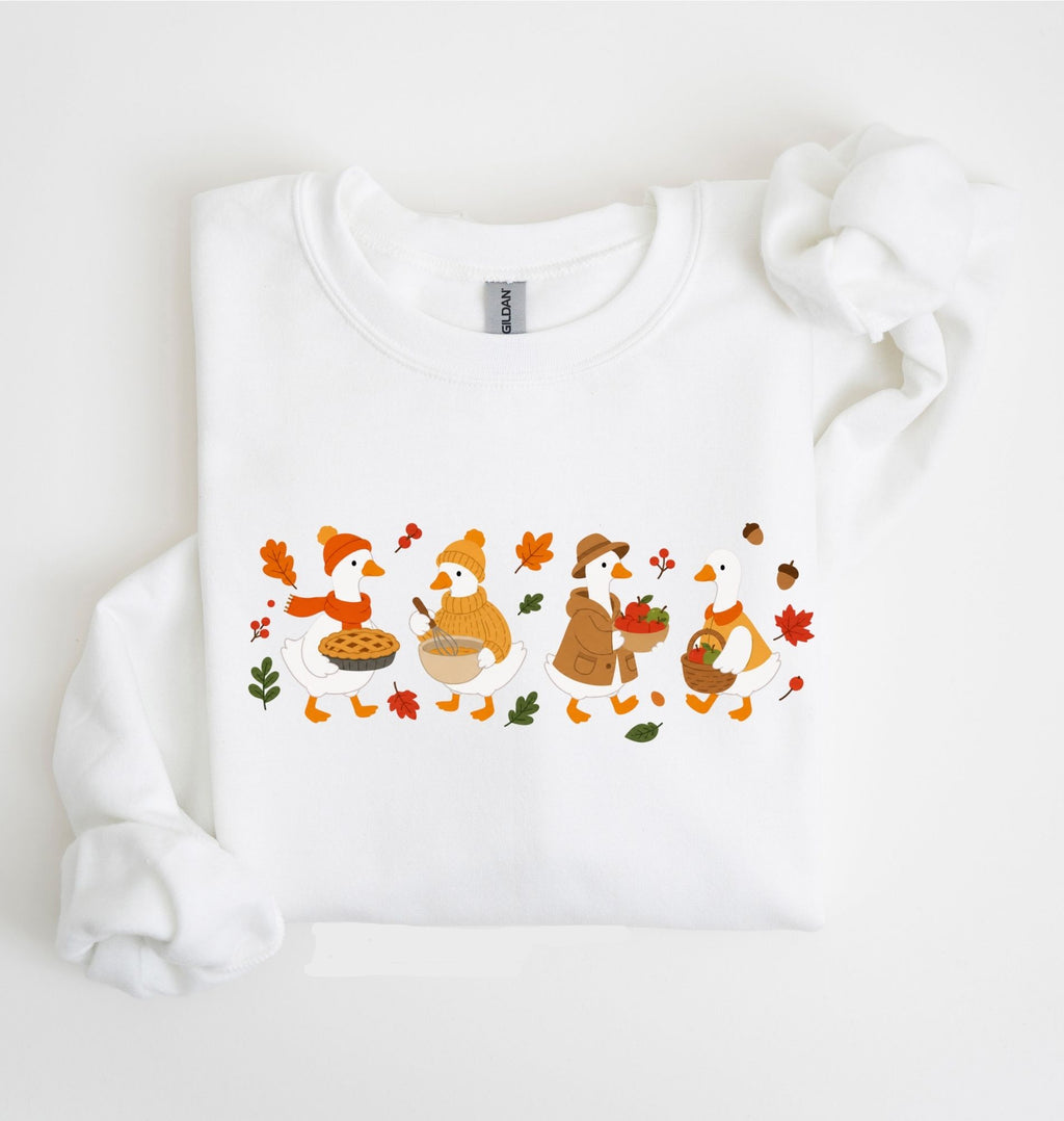 Flat lay of a white fall sweatshirt featuring four geese baking pie, mixing ingredients and gathering apples on a white background