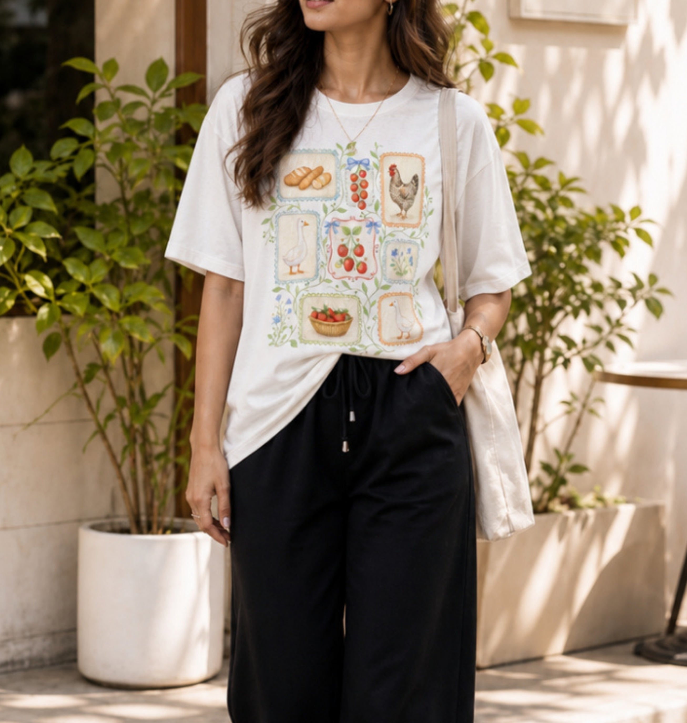 A woman wearing a white graphic t-shirt with a colorful farm-themed print and black drawstring sweatpants, standing in an outdoor patio setting with potted plants.