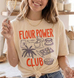 A woman in a kitchen wearing a cream-colored graphic t-shirt with orange and dark purple text that reads 'FLOUR POWER CLUB' along with illustrations of baking items. She is holding a whisk covered in flour.