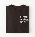 Flat lay of a brown flour water salt shirt black minimalist sourdough baking t shirt bread lover gift simple design on a white background 