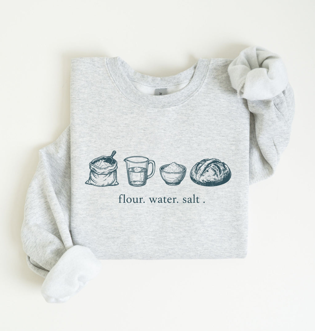 A light heather gray sweatshirt with long sleeves, laid flat on a white background. The front of the sweatshirt features a horizontal graphic illustration of baking ingredients, including a bowl, flour sack, butter, sugar jar, vanilla bottle, eggs, and a measuring cup.
