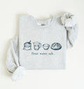 A light heather gray sweatshirt with long sleeves, laid flat on a white background. The front of the sweatshirt features a horizontal graphic illustration of baking ingredients, including a bowl, flour sack, butter, sugar jar, vanilla bottle, eggs, and a measuring cup.