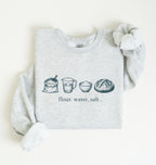 A light heather gray sweatshirt with long sleeves, laid flat on a white background. The front of the sweatshirt features a horizontal graphic illustration of baking ingredients, including a bowl, flour sack, butter, sugar jar, vanilla bottle, eggs, and a measuring cup.