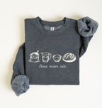 A dark heather gray crewneck sweatshirt with rolled cuffs, featuring a graphic print of baking ingredients (bowl, flour sack, butter, sugar jar, eggs, and measuring cup) across the chest, laid flat on a light cream background.