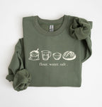 An olive green long-sleeve sweatshirt with a graphic of baking ingredients (milk, sunflower butter, sugar, eggs, vanilla, flour) on the chest, styled with a large scrunchie on the shoulder and a matching scrunchie on the sleeve, against a plain white background.