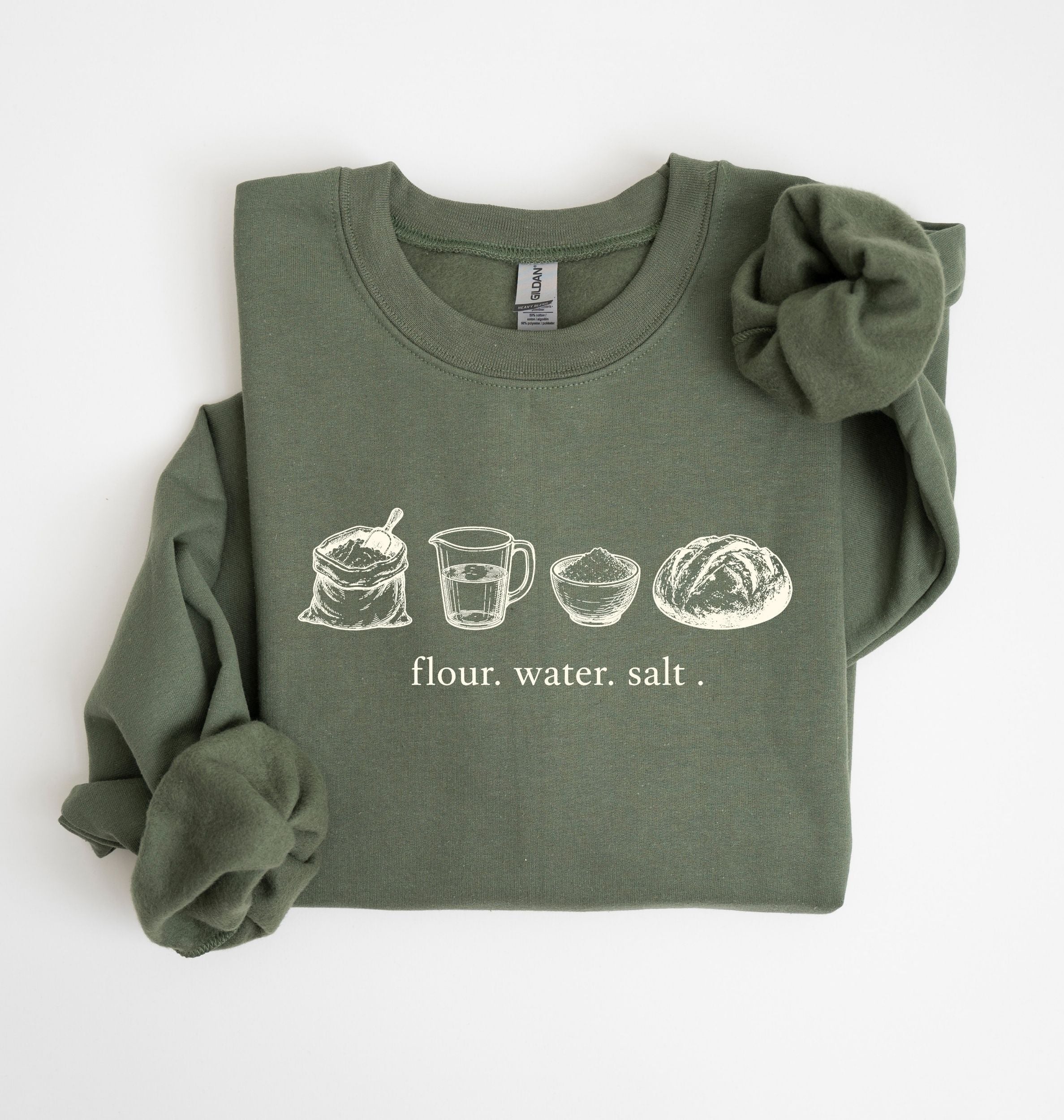 An olive green long-sleeve sweatshirt with a graphic of baking ingredients (milk, sunflower butter, sugar, eggs, vanilla, flour) on the chest, styled with a large scrunchie on the shoulder and a matching scrunchie on the sleeve, against a plain white background.