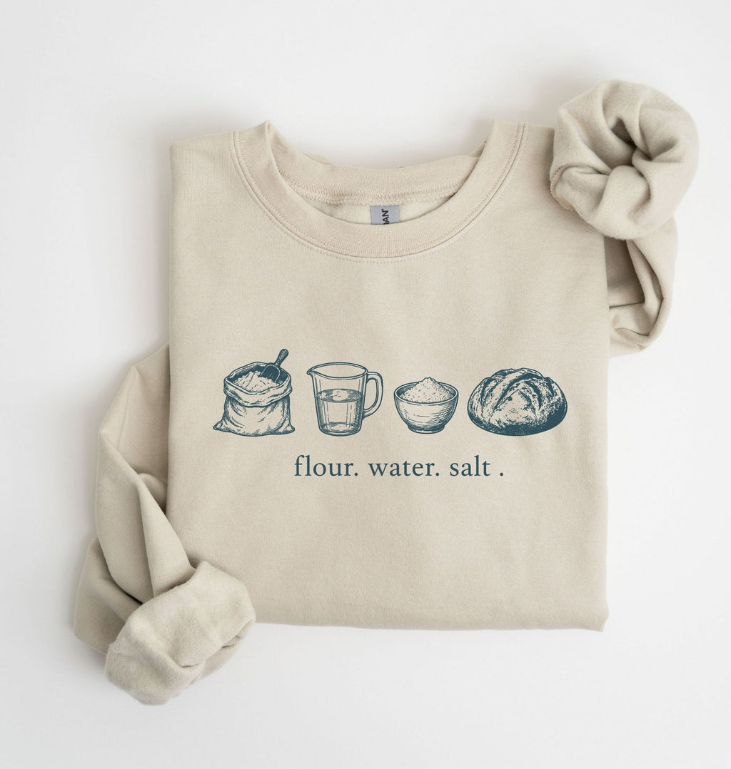 A beige crewneck sweatshirt with a graphic of baking ingredients (bowl, flour, butter, sugar, vanilla, eggs, measuring cup) printed on the chest, styled flat on a white background with a matching fabric scrunchie.