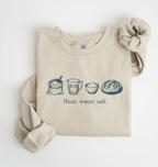 A beige crewneck sweatshirt with a graphic of baking ingredients (bowl, flour, butter, sugar, vanilla, eggs, measuring cup) printed on the chest, styled flat on a white background with a matching fabric scrunchie.
