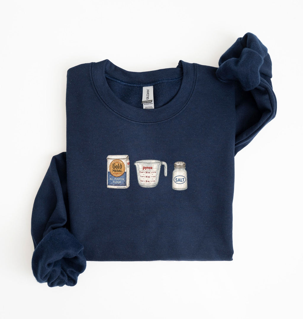 Flat lay of a navy blue flour water salt sweatshirt with flour bag measuring cup and salt shaker for bread bakers on a white background 