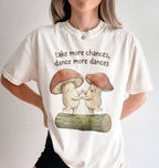 Flat lay of a Pepper ivory t-shirt with two dancing mushrooms and the phrase take more chances dance more dances, whimsical cottagecore gift tee on a white background 