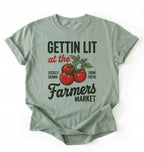 Flat lay of a bay t-shirt featuring tomatoes and text reading “Gettin Lit at the Farmers Market Locally Grown Farm Fresh.” Funny farmers market shirt pun perfect gift for gardeners homesteaders and vegetable lovers on a white background 