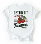 Flat lay of a white t-shirt featuring tomatoes and text reading “Gettin Lit at the Farmers Market Locally Grown Farm Fresh.” Funny farmers market shirt pun perfect gift for gardeners homesteaders and vegetable lovers on a white background 