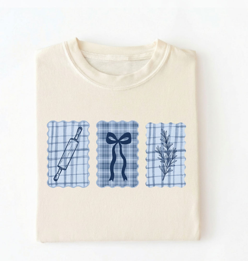 Flat lay of an ivory Cottagecore baking shirt with blue gingham pattern and baking icons perfect gift for bakers and vintage kitchen lovers on a white background 