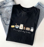 A black graphic t-shirt with a 'It's a good day to bake' illustration, styled flat-lay on top of light-wash denim jeans against a white background.