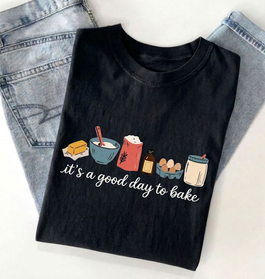 A black graphic t-shirt with a 'it's a good day to bake' illustration, styled flat-lay on top of light blue jeans against a white background.