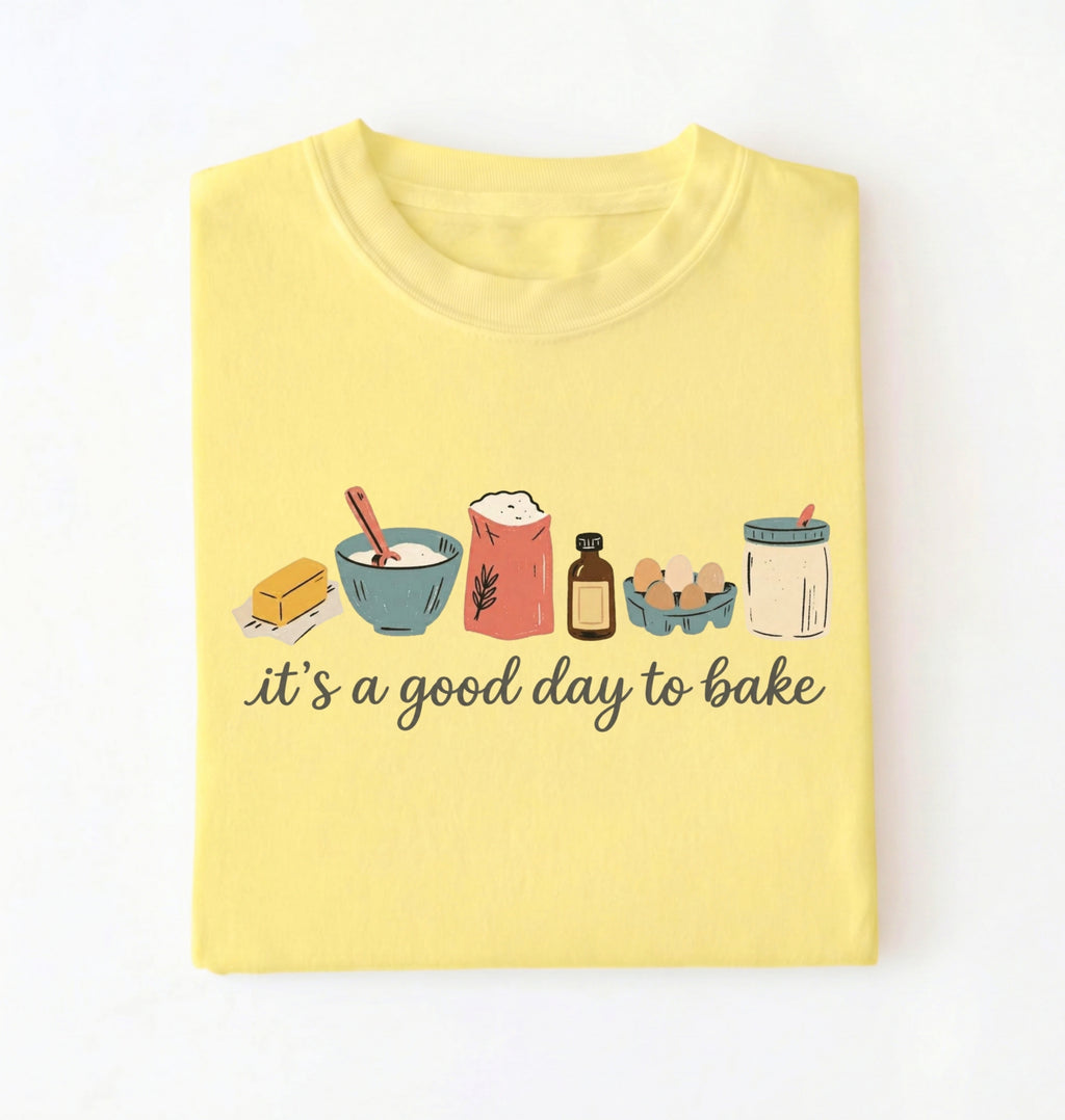 A light yellow folded t-shirt on a white background, featuring a graphic of baking ingredients and the text 'it's a good day to bake' in black script.