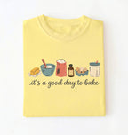 A light yellow folded t-shirt on a white background, featuring a graphic of baking ingredients and the text 'it's a good day to bake' in black script.