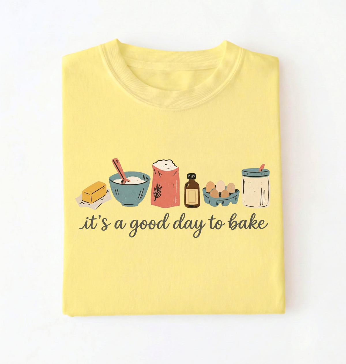 A light yellow folded t-shirt on a white background, featuring a graphic of baking ingredients and the text 'it's a good day to bake' in black script.