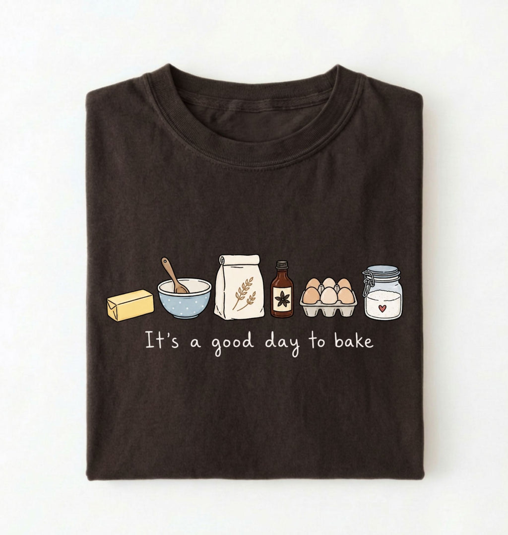 A dark brown folded t-shirt with a graphic of baking ingredients and the text 'It's a good day to bake' on a white background.
