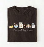 A dark brown folded t-shirt with a graphic of baking ingredients and the text 'It's a good day to bake' on a white background.
