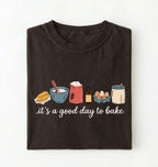 A dark brown folded t-shirt with a graphic of baking ingredients and the text 'it's a good day to bake' on a white background.