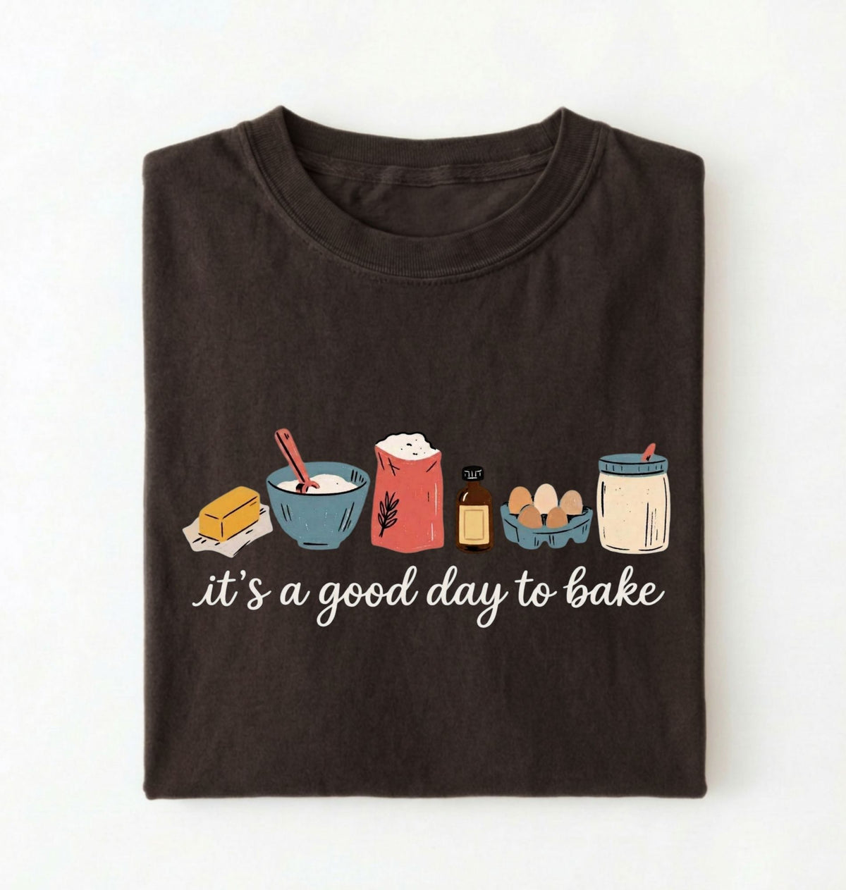A dark brown folded t-shirt with a graphic of baking ingredients and the text 'it's a good day to bake' on a white background.