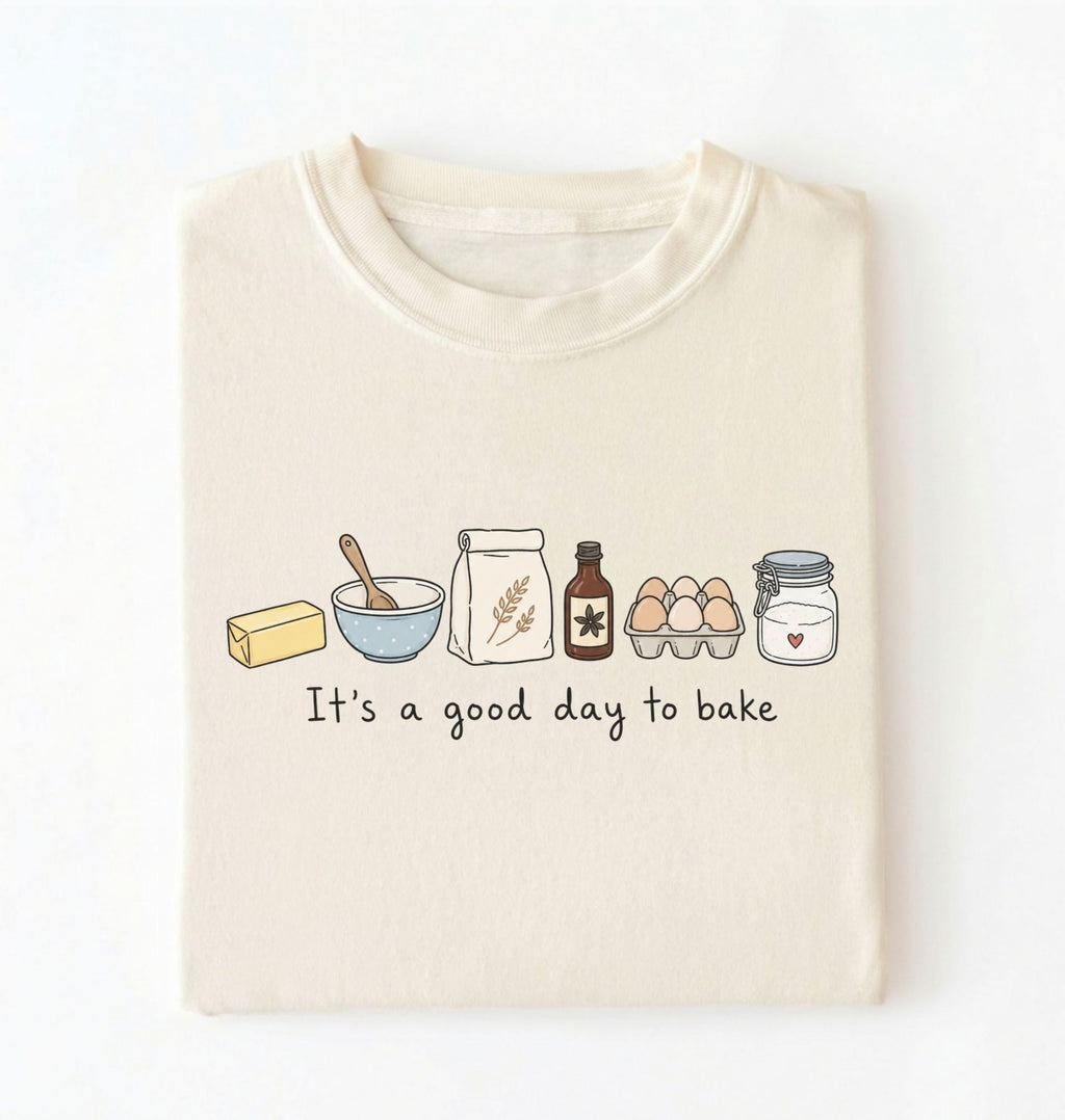 A folded cream-colored t-shirt with a graphic of baking ingredients and the text 'It's a good day to bake' printed on the front, displayed against a clean white background.