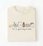 A folded cream-colored t-shirt with a graphic of baking ingredients and the text 'It's a good day to bake' printed on the front, displayed against a clean white background.