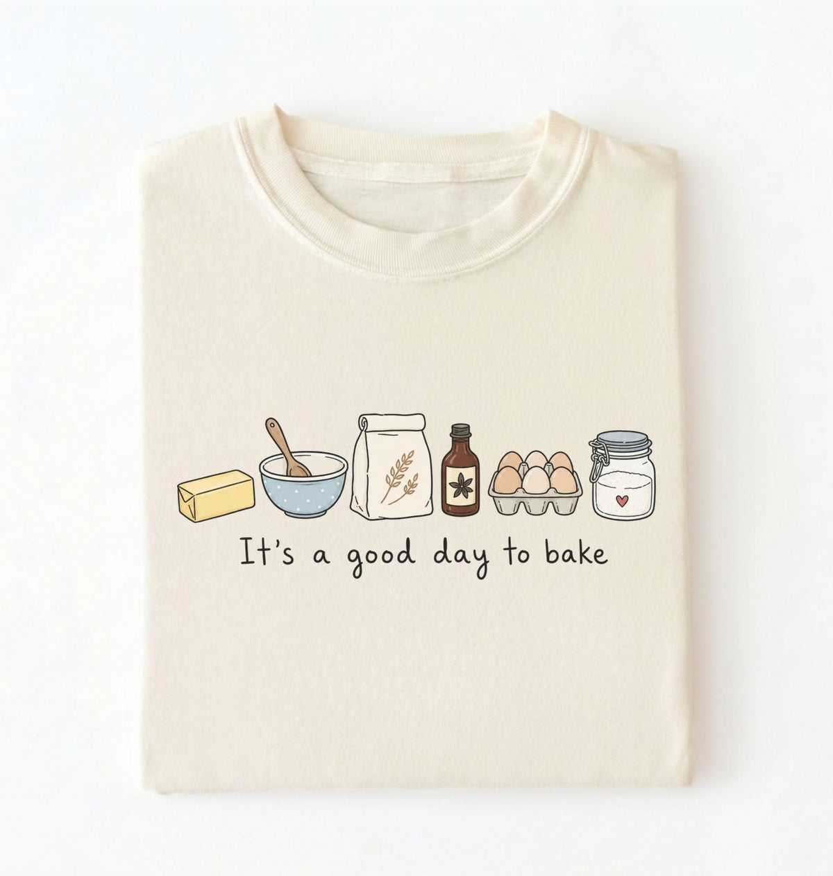 A folded cream-colored t-shirt with a graphic of baking ingredients and the text 'It's a good day to bake' printed on the front, displayed against a clean white background.