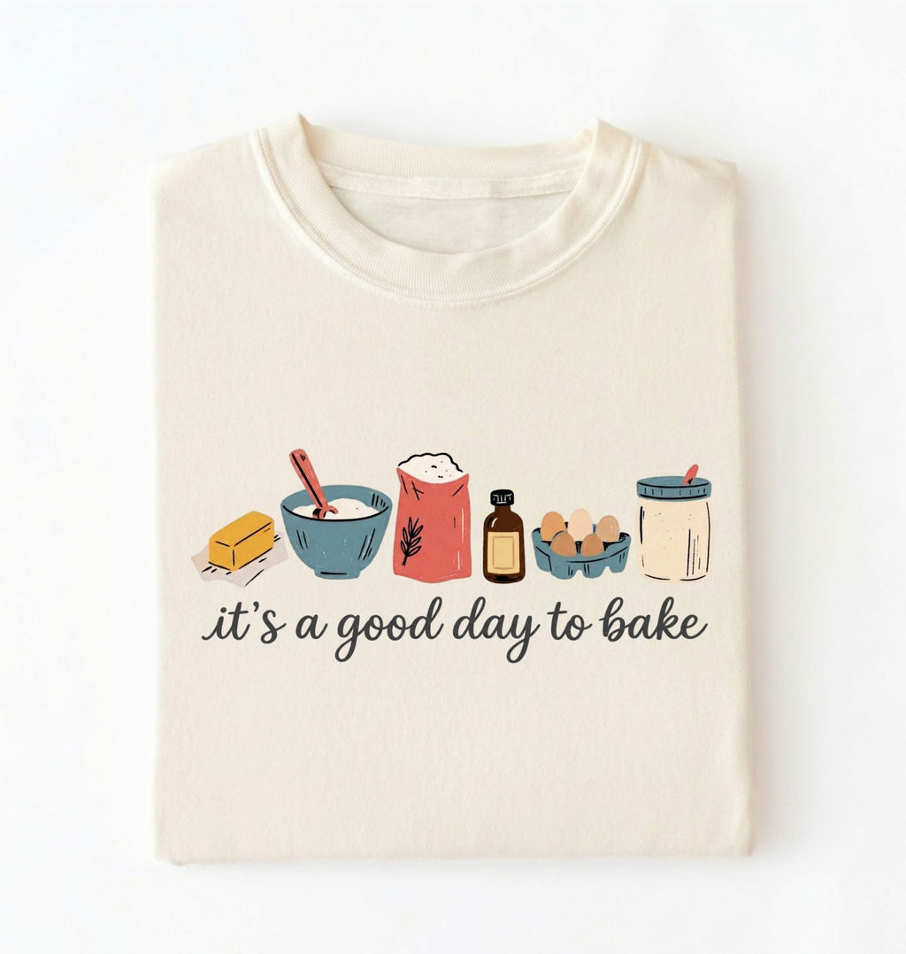 A cream-colored t-shirt with a graphic illustration of baking ingredients and the text 'it's a good day to bake' printed on the front, displayed flat against a white background.