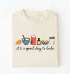 A cream-colored t-shirt with a graphic illustration of baking ingredients and the text 'it's a good day to bake' printed on the front, displayed flat against a white background.