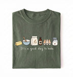 An olive green folded t-shirt with a graphic of baking ingredients and the text 'It's a good day to bake' on a white background.