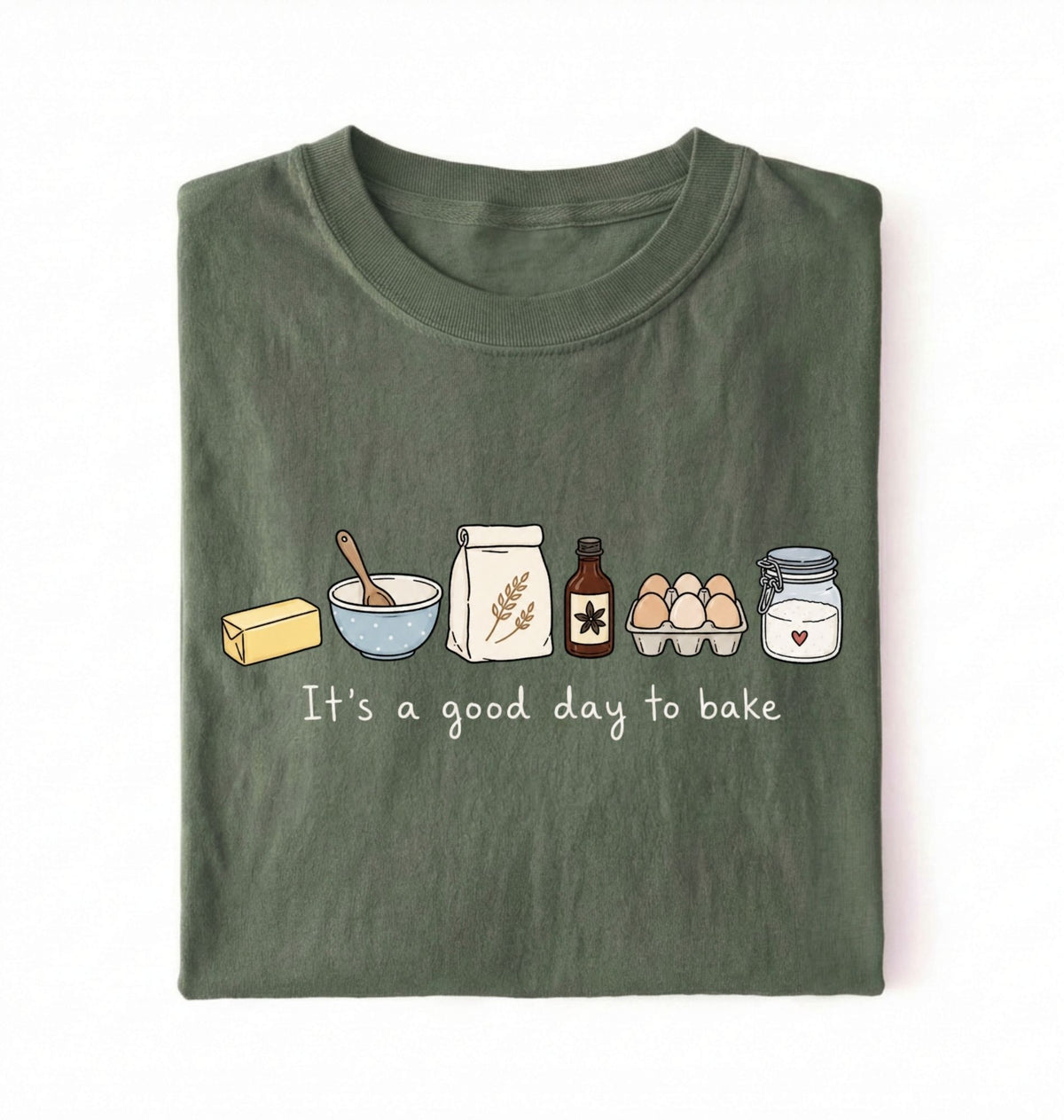 An olive green folded t-shirt with a graphic of baking ingredients and the text 'It's a good day to bake' on a white background.