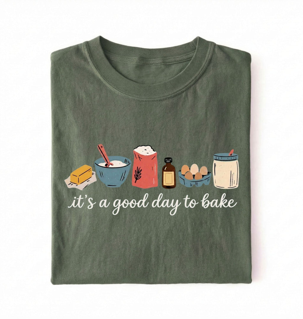 A folded dark olive green t-shirt on a white background, featuring a graphic of baking ingredients and the text 'it's a good day to bake' in white script.