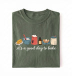 A folded dark olive green t-shirt on a white background, featuring a graphic of baking ingredients and the text 'it's a good day to bake' in white script.