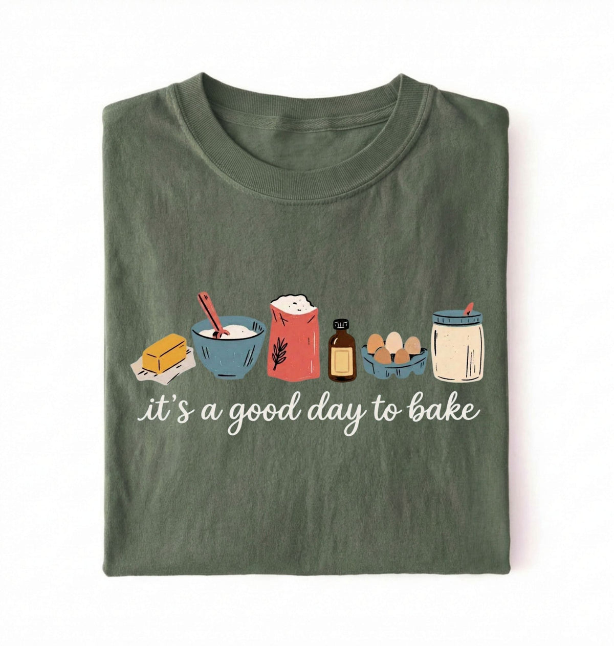 A folded dark olive green t-shirt on a white background, featuring a graphic of baking ingredients and the text 'it's a good day to bake' in white script.