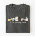 A dark gray folded t-shirt with a graphic of baking ingredients and the text 'It's a good day to bake' on a white background.