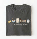 A dark gray folded t-shirt with a graphic of baking ingredients and the text 'It's a good day to bake' on a white background.