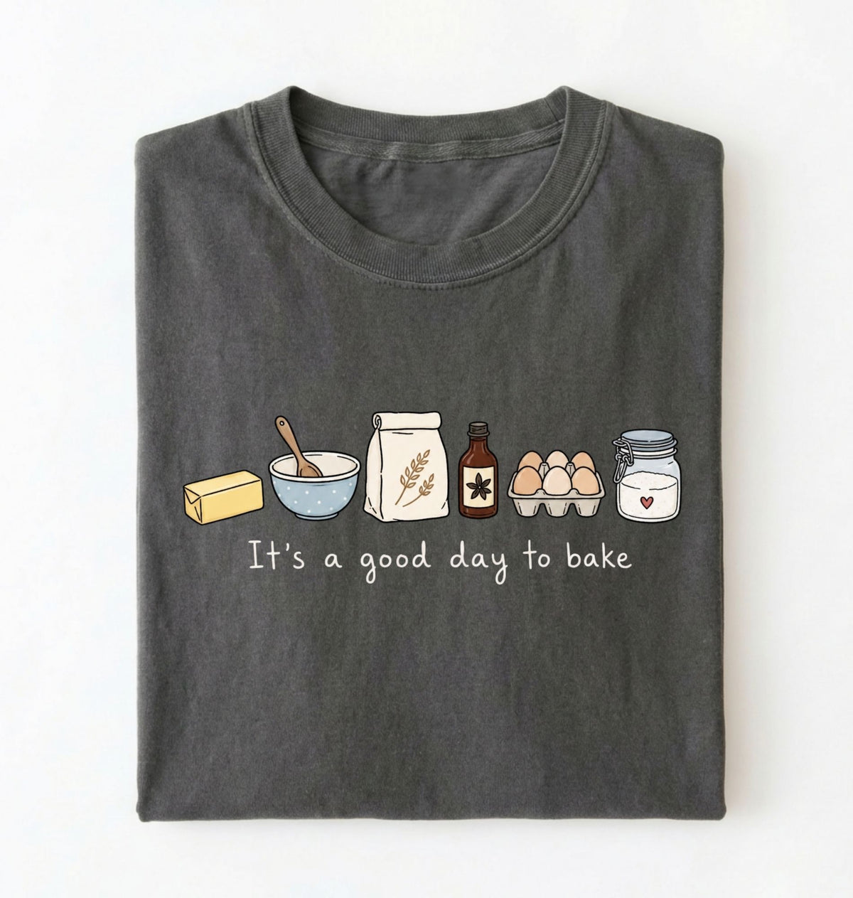 A dark gray folded t-shirt with a graphic of baking ingredients and the text 'It's a good day to bake' on a white background.