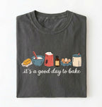 A dark gray folded t-shirt with a graphic of baking ingredients and the text 'it's a good day to bake' on a white background.