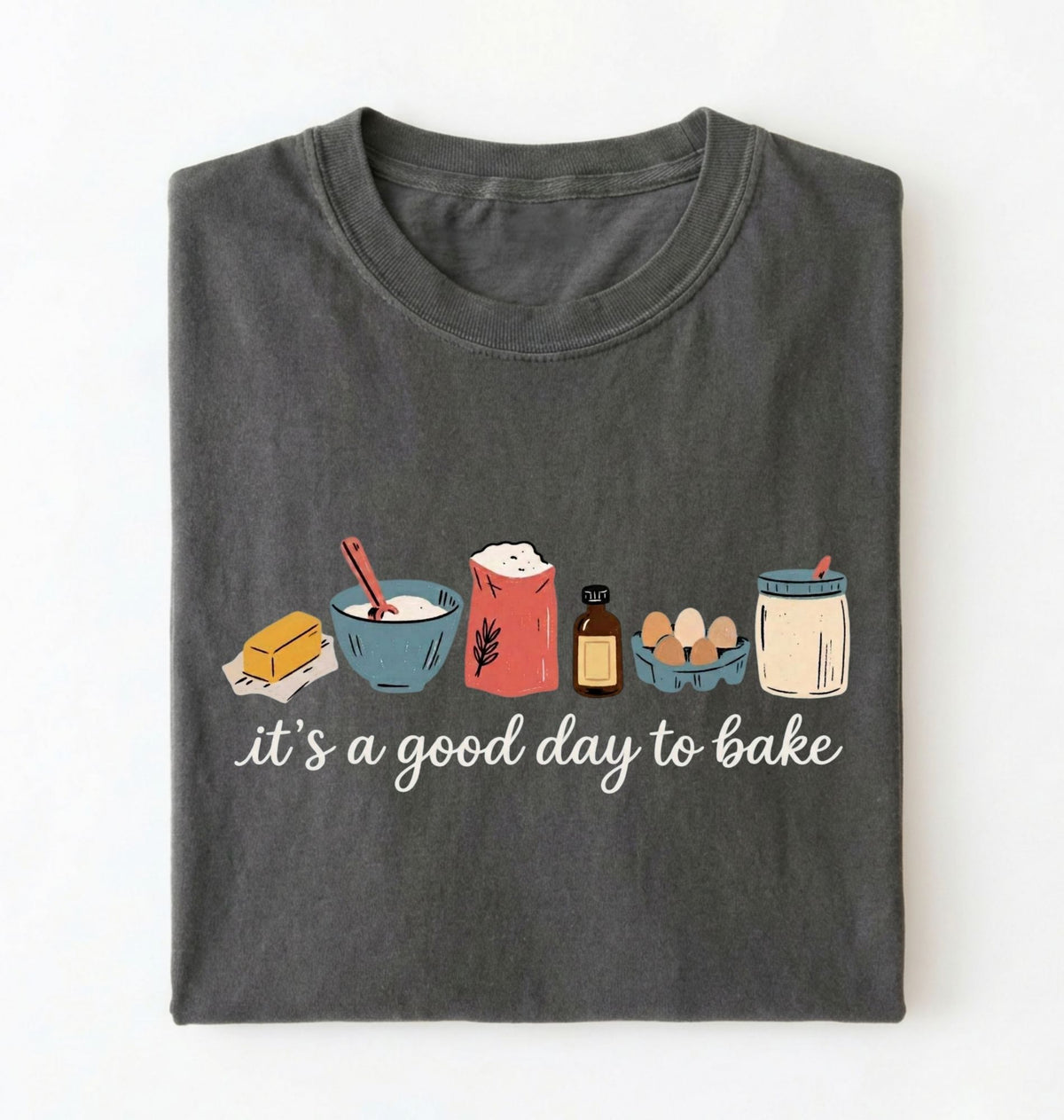 A dark gray folded t-shirt with a graphic of baking ingredients and the text 'it's a good day to bake' on a white background.