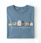 A folded slate blue t-shirt with a cute baking-themed graphic on the front. The graphic shows illustrated ingredients (butter, bowl, flour, vanilla, eggs, sugar) and the text 'It's a good day to bake' in white script.