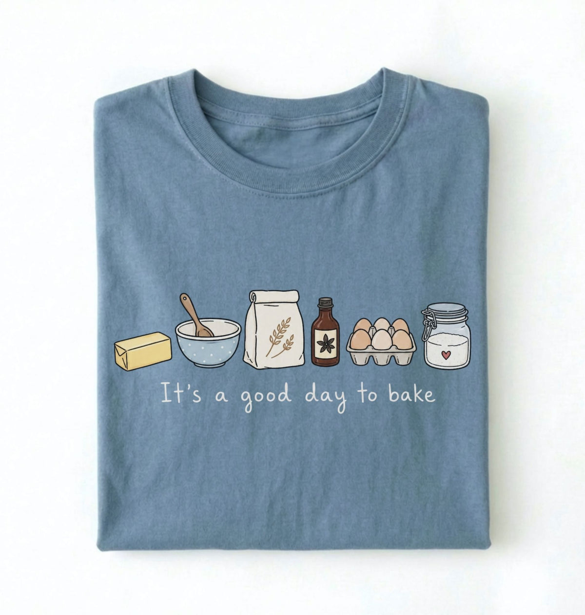 A folded slate blue t-shirt with a cute baking-themed graphic on the front. The graphic shows illustrated ingredients (butter, bowl, flour, vanilla, eggs, sugar) and the text 'It's a good day to bake' in white script.