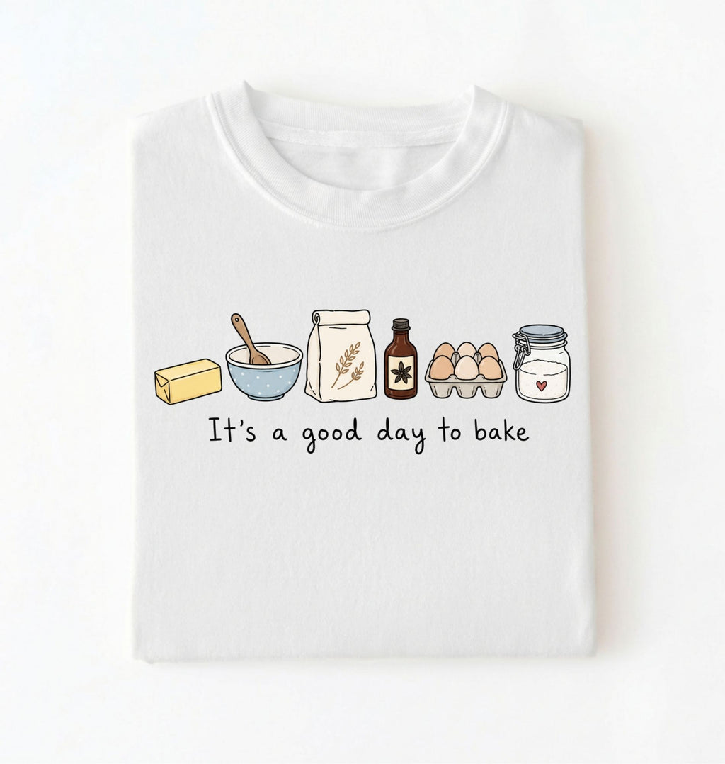 A folded white t-shirt with a graphic of baking ingredients and the text 'It's a good day to bake' printed on the front, displayed against a clean white background.