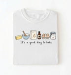 A folded white t-shirt with a graphic of baking ingredients and the text 'It's a good day to bake' printed on the front, displayed against a clean white background.