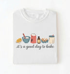 A white folded t-shirt with a graphic illustration of baking ingredients and the text 'it's a good day to bake' printed on the front, set against a plain white background.
