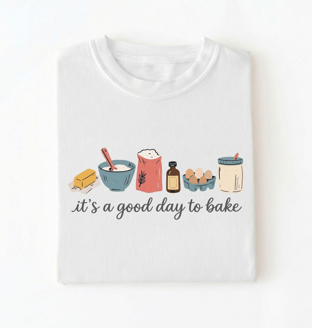A white folded t-shirt with a graphic illustration of baking ingredients and the text 'it's a good day to bake' printed on the front, set against a plain white background.