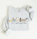 A light heather gray long-sleeve sweatshirt with a graphic of baking ingredients and the text 'It's a good day to bake' on the front, styled flat on a light cream background.