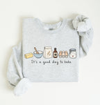 A light heather gray long-sleeve sweatshirt with a graphic of baking ingredients and the text 'It's a good day to bake' on the front, styled flat on a light cream background.